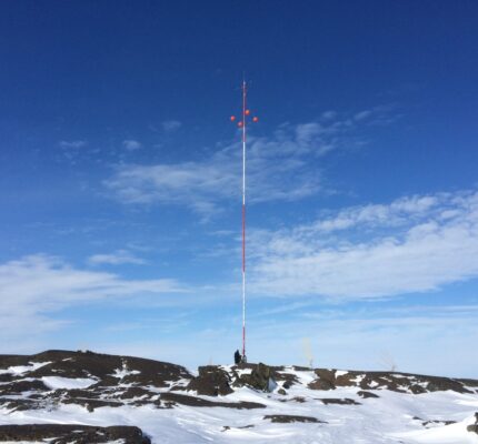 Tall meteorological tower with red and white stripes on a snow-covered, rocky Antarctic landscape under a clear blue sky.
