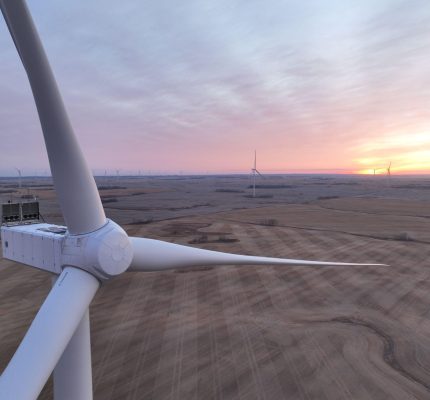 Gros plan sur la nacelle et les pales d’une éolienne au-dessus d’un vaste champ moissonné au lever ou au coucher du soleil, avec d’autres éoliennes visibles à l’horizon.