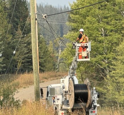 Worker in a high‑visibility safety vest and hard hat stands on an elevated bucket attached to a utility truck, installing broadband infrastructure on overhead lines at a roadside utility pole. The truck is parked on a narrow rural road with tall dry grass and dense green trees on both sides. A large spool of cable is mounted on the truck, with forested hills in the background.