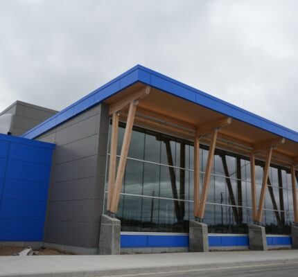 Exterior of the Thompson regional airport terminal featuring bright blue and dark grey cladding, large floor-to-ceiling windows, and a wood-paneled overhang supported by tall timber columns.