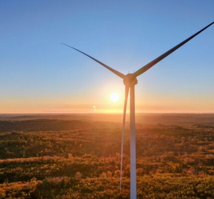 A towering wind turbine stands silhouetted against a bright sunset over a vast, forested landscape at the Benjamins Mill Wind completed site.