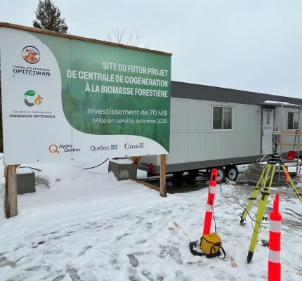 Un panneau annonçant la future centrale de cogénération à la biomasse forestière à Opitciwan est installé à côté d'une remorque de chantier temporaire dans un paysage enneigé.