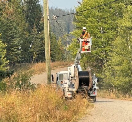 Worker in a high‑visibility safety vest and hard hat stands on an elevated bucket attached to a utility truck, installing broadband infrastructure on overhead lines at a roadside utility pole. The truck is parked on a narrow rural road with tall dry grass and dense green trees on both sides. A large spool of cable is mounted on the truck, with forested hills in the background.