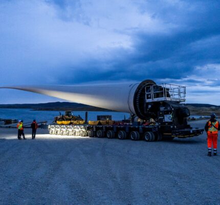 Industrial construction site in a rocky Arctic landscape, with cranes, buildings and equipment beneath a cliff. In the foreground, a wind turbine blade is transported along a gravel road under an overcast sky.