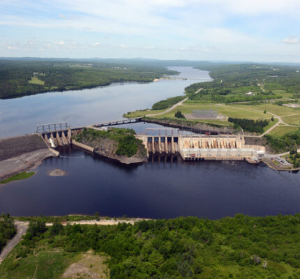 Aerial view of the Mactaquac Generating Station and reservoir on the Saint John River, surrounded by forested landscape and electrical infrastructure.
