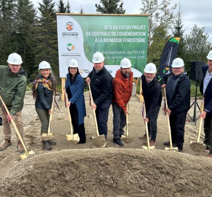 Un groupe de huit personnes avec des casques de chantier participe à la cérémonie d'inauguration du début des travaux des futures installations de la centrale de cogénération à la biomasse forestière à Opitciwan, tenant des pelles devant un grand panneau.