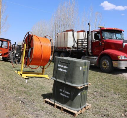 Bright day scene with a red truck, broadband construction equipment, including a fibre cable spool and service boxes, along a rural roadside. Leafless trees and a blue sky are in the background.