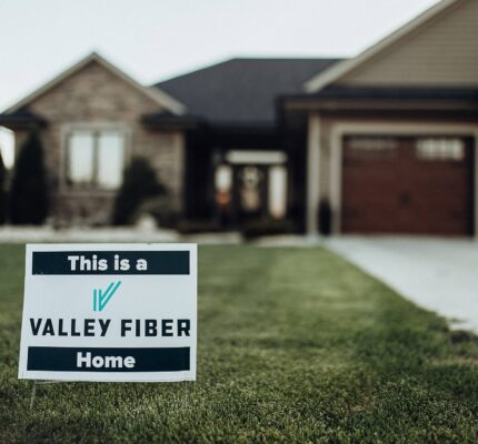 A sign on a lush green lawn reads “This is a Valley Fiber Home,” in front of a large, modern brick house with a driveway and garage.