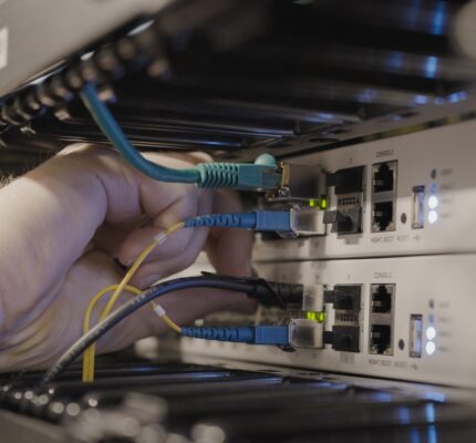 Hand connecting blue and yellow network cables to switches inside a server rack, with indicator lights illuminated.