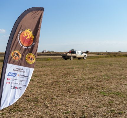 A feather flag for Wicehtowak Solar Ltd., mentioning the partnership with the CIB and other organizations, stands in the foreground of a dry, open field with a white pickup truck and construction equipment visible in the distance under a clear blue sky.