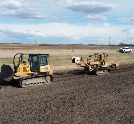 Yellow construction equipment install fibre optic cables farmland adjacent to a rural road.