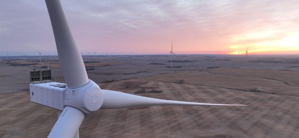 Gros plan sur la nacelle et les pales d’une éolienne au-dessus d’un vaste champ moissonné au lever ou au coucher du soleil, avec d’autres éoliennes visibles à l’horizon.