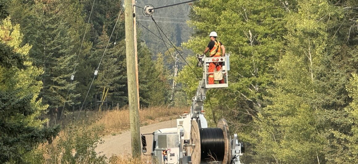 Worker in a high‑visibility safety vest and hard hat stands on an elevated bucket attached to a utility truck, installing broadband infrastructure on overhead lines at a roadside utility pole. The truck is parked on a narrow rural road with tall dry grass and dense green trees on both sides. A large spool of cable is mounted on the truck, with forested hills in the background.
