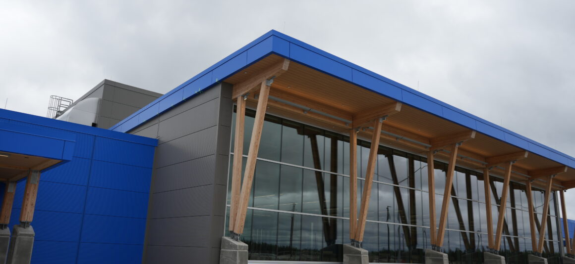 Exterior of the Thompson regional airport terminal featuring bright blue and dark grey cladding, large floor-to-ceiling windows, and a wood-paneled overhang supported by tall timber columns.