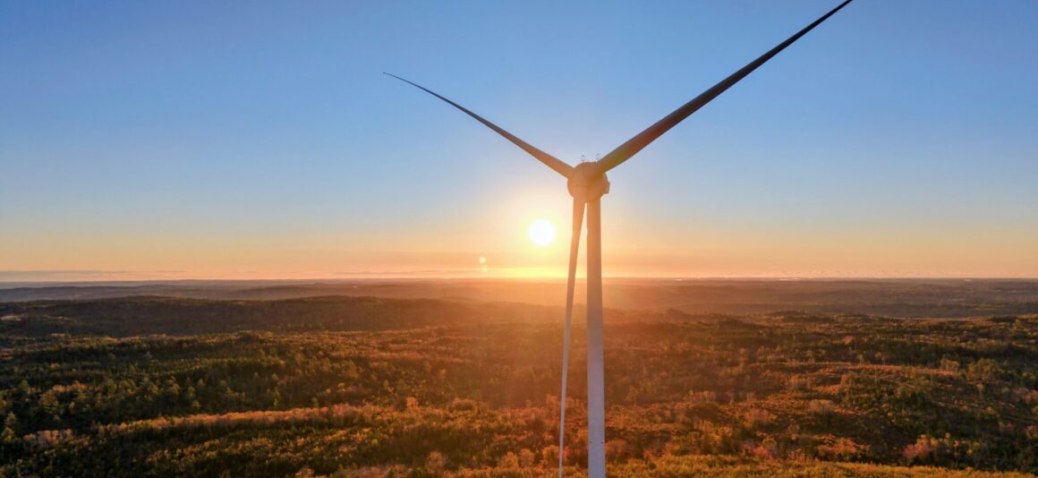 A towering wind turbine stands silhouetted against a bright sunset over a vast, forested landscape at the Benjamins Mill Wind completed site.