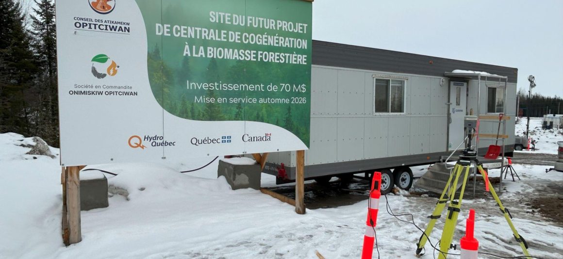 Un panneau annonçant la future centrale de cogénération à la biomasse forestière à Opitciwan est installé à côté d'une remorque de chantier temporaire dans un paysage enneigé.