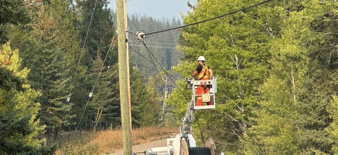 Worker in a high‑visibility safety vest and hard hat stands on an elevated bucket attached to a utility truck, installing broadband infrastructure on overhead lines at a roadside utility pole. The truck is parked on a narrow rural road with tall dry grass and dense green trees on both sides. A large spool of cable is mounted on the truck, with forested hills in the background.
