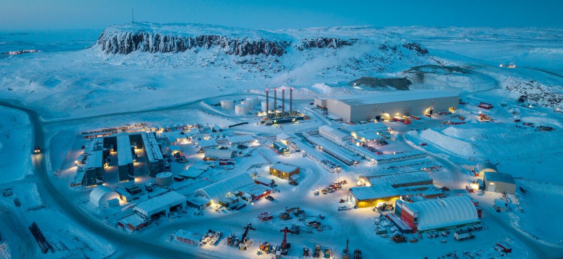 Aerial view of the Hope Bay Mine and processing site in a snow-covered Arctic landscape at dusk. Illuminated buildings, storage areas, roads and heavy equipment are spread across the site.