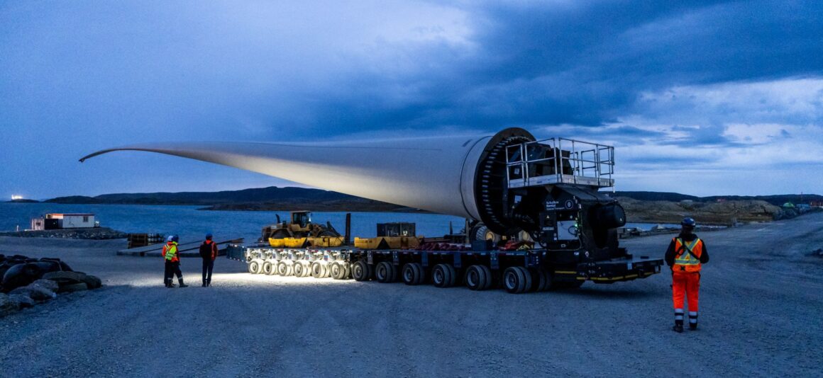Industrial construction site in a rocky Arctic landscape, with cranes, buildings and equipment beneath a cliff. In the foreground, a wind turbine blade is transported along a gravel road under an overcast sky.