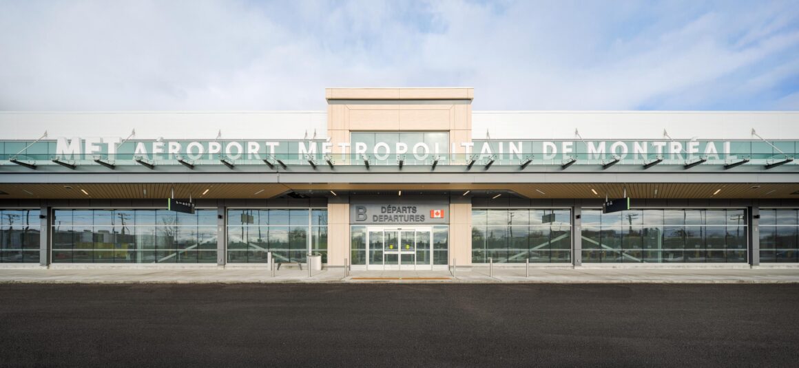 Vue frontale de l’entrée de l’aérogare pour vols intérieurs de l’aéroport métropolitain de Montréal (YHU), avec sa façade en verre, ses portes d’entrée coulissantes et le nom du bâtiment inscrit sur l’auvent extérieur supérieur.