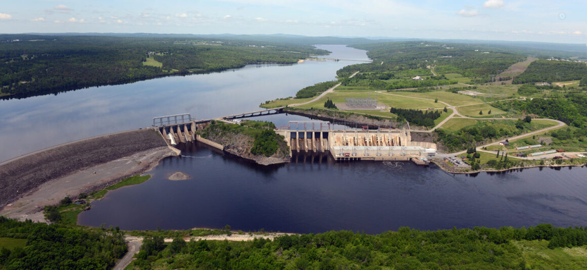 Aerial view of the Mactaquac Generating Station and reservoir on the Saint John River, surrounded by forested landscape and electrical infrastructure.