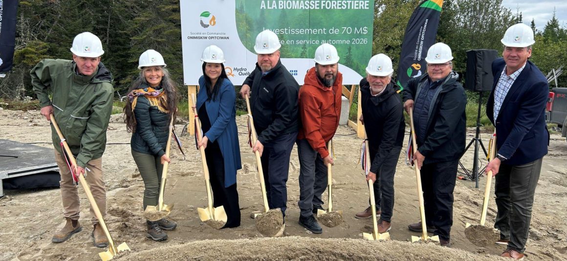 A group of eight people wearing hard hats participate in the groundbreaking ceremony for the future forest biomass cogeneration plant in Opitciwan, holding shovels in front of a large sign.