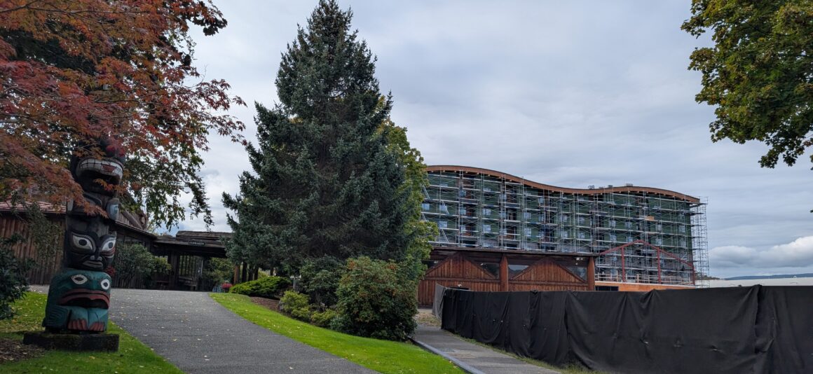 Landscaped pathway leading toward a large building under construction, with scaffolding covering the exterior. Trees with autumn foliage and a carved totem pole are visible in the foreground.