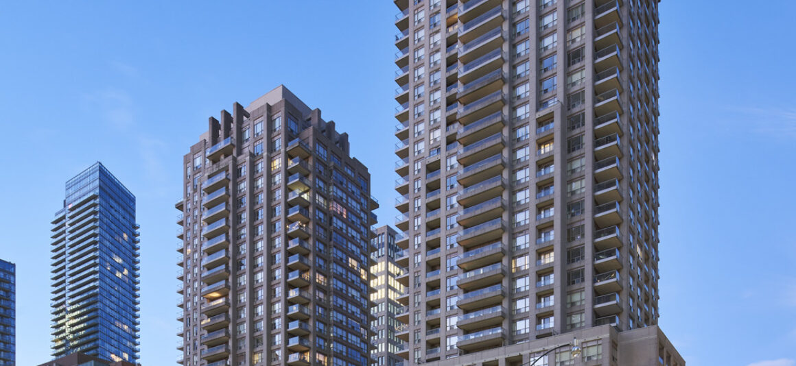 Les tours du Bay Club. Des tours résidentielles de grande hauteur s’élèvent au-dessus d’une rue de la ville au crépuscule, avec des balcons et des fenêtres illuminés sur un ciel bleu clair. Au premier plan, une zone de parc paysager présente des arbres avec un feuillage d’automne et une structure sculpturale moderne, tandis que des lampadaires et des immeubles mi-hauteur environnants encadrent la scène urbaine.