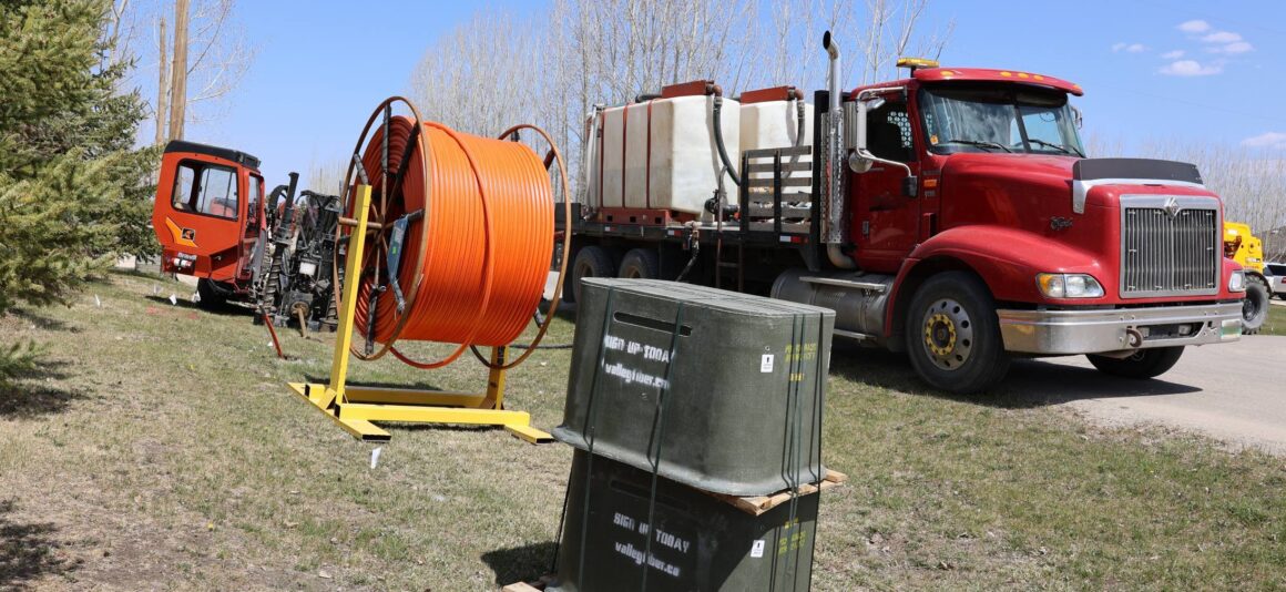 Bright day scene with a red truck, broadband construction equipment, including a fibre cable spool and service boxes, along a rural roadside. Leafless trees and a blue sky are in the background.