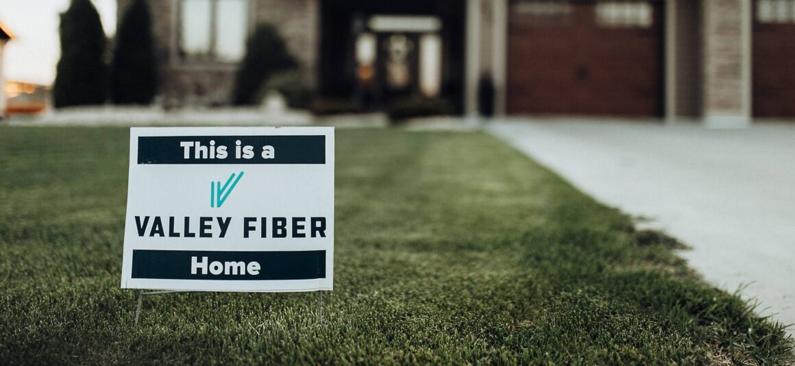 A sign on a lush green lawn reads “This is a Valley Fiber Home,” in front of a large, modern brick house with a driveway and garage.