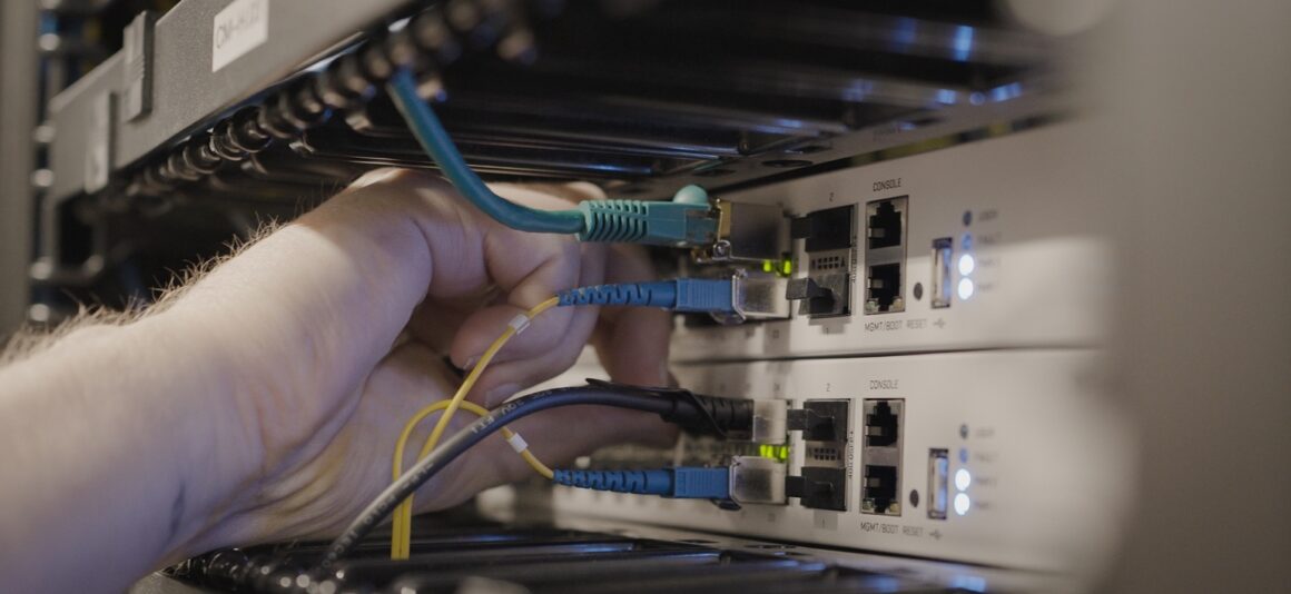 Hand connecting blue and yellow network cables to switches inside a server rack, with indicator lights illuminated.