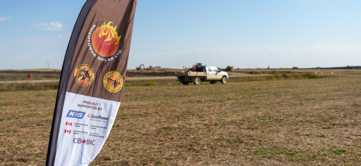 A feather flag for Wicehtowak Solar Ltd., mentioning the partnership with the CIB and other organizations, stands in the foreground of a dry, open field with a white pickup truck and construction equipment visible in the distance under a clear blue sky.