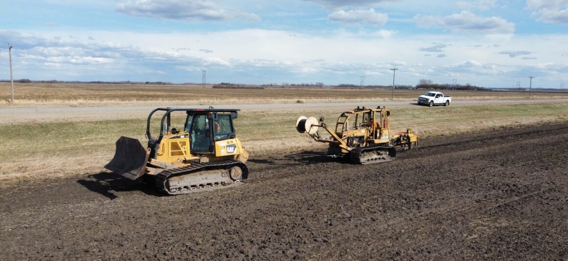 Yellow construction equipment install fibre optic cables farmland adjacent to a rural road.