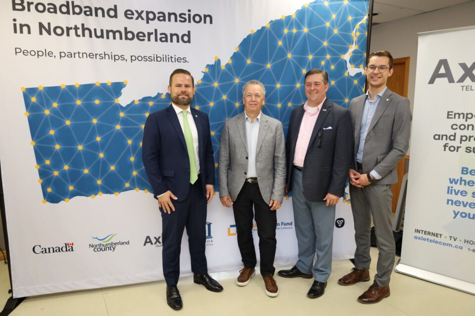 Four provincial Ministers in suits stand before a backdrop displaying broadband expansion in Northumberland County, Canada.