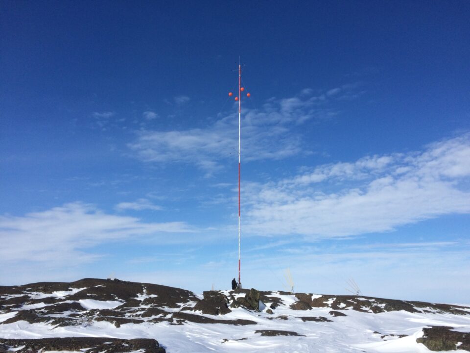 Tall meteorological tower with red and white stripes on a snow-covered, rocky Antarctic landscape under a clear blue sky.