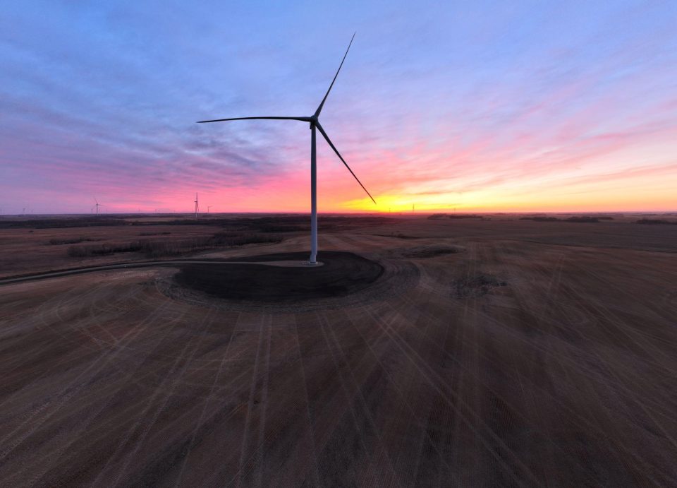 Une éolienne isolée se dresse dans un vaste champ moissonné, avec en toile de fond un ciel spectaculaire aux teintes roses, violettes et orangées.