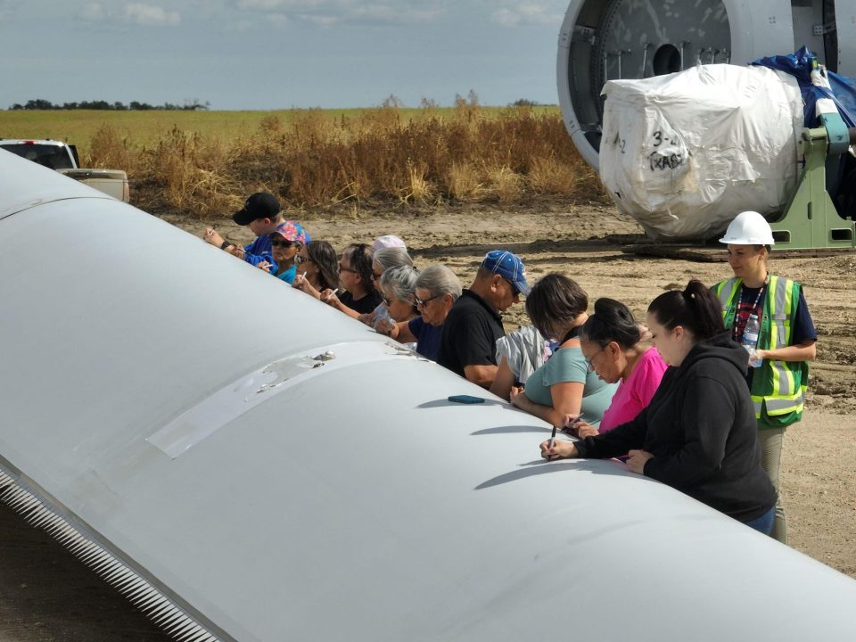 Un groupe des aînés de la communauté se rassemble à l’extérieur pour signer une grande pale d’éolienne grise, sous la supervision d’un ouvrier portant un casque blanc et un gilet de sécurité.