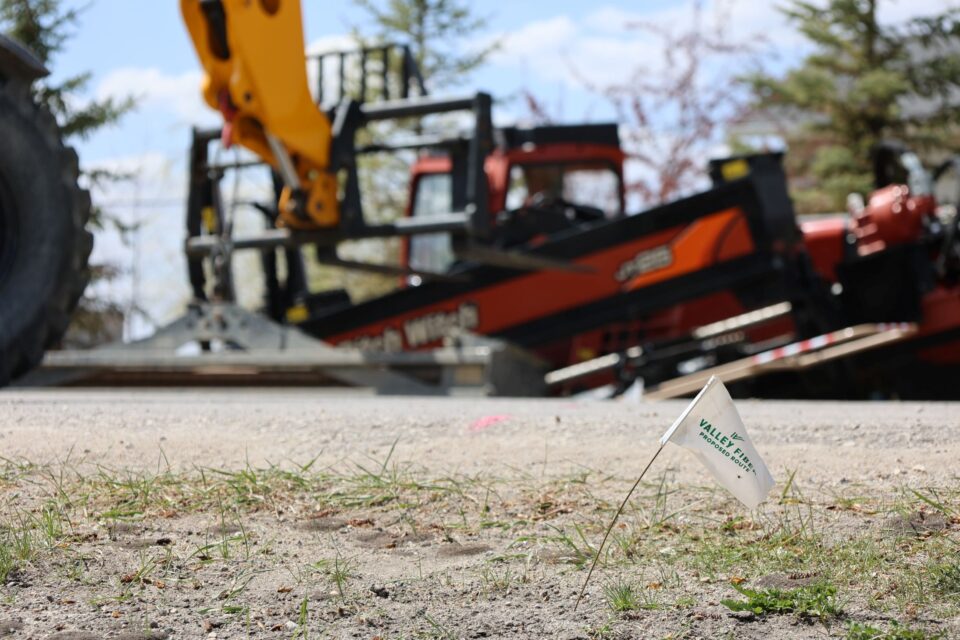 A small white Valley Fiber utility flag with green text stands on a dirt road, marking underground infrastructure near active broadband construction equipment, with blurred construction machinery in the background.