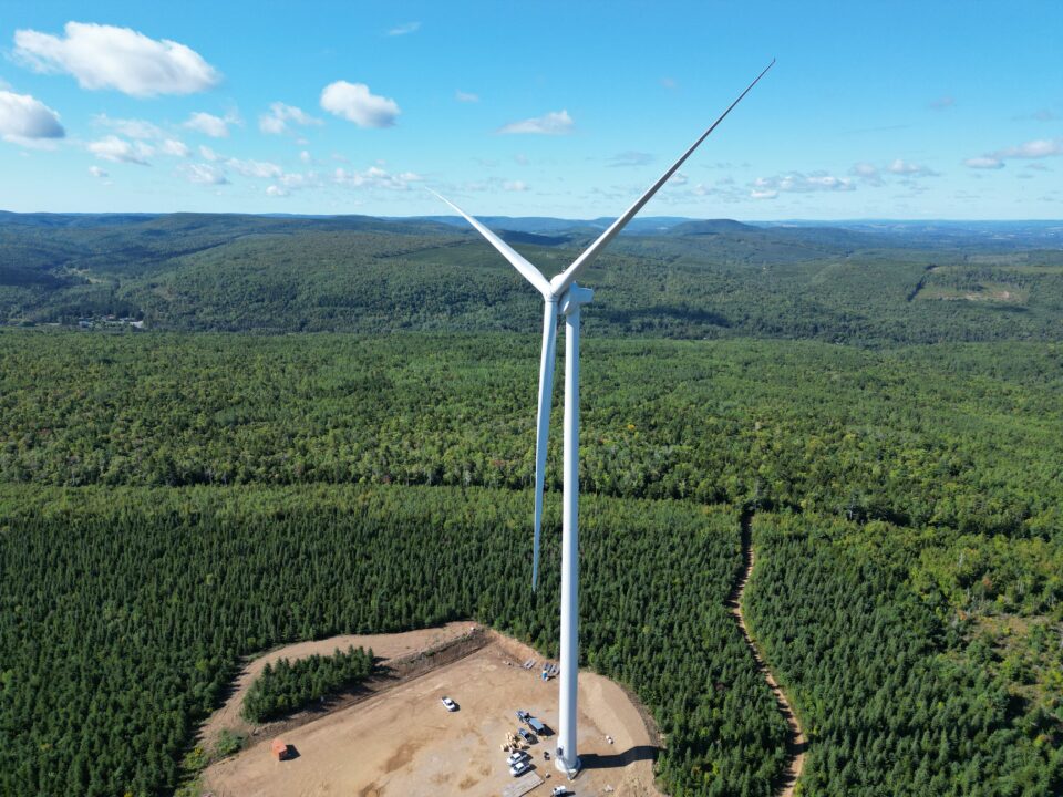 Vue aérienne d'une éolienne au-dessus d'un paysage forestier. L'éolienne se dresse dans une zone de construction dégagée entourée d'arbres.