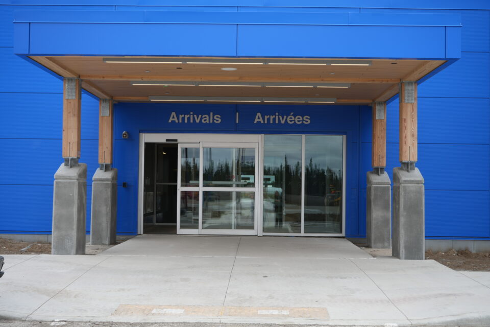 Exterior entrance to a modern building with bright blue cladding and a wood-paneled canopy supported by timber columns. Glass sliding doors sit beneath bilingual signage reading