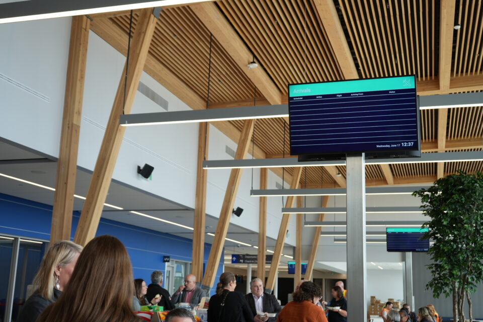 Interior of a modern airport terminal with high wood-slat ceilings, exposed timber supports and suspended linear lighting. Passengers gather beneath digital arrivals screens, with seating and a large indoor plant visible in the background.