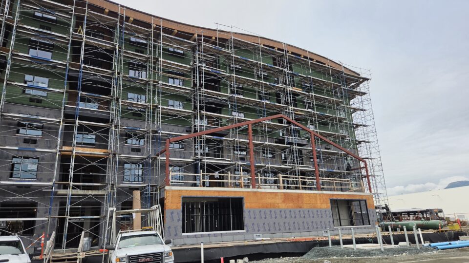 Exterior view of a multi-storey building under construction, surrounded by scaffolding, with exposed structural elements and construction vehicles in the foreground.