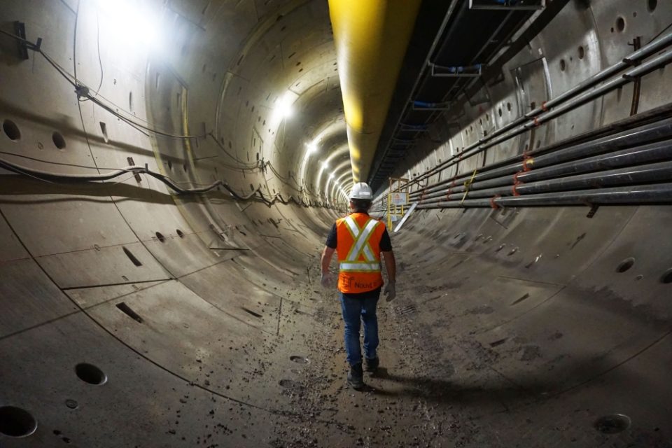 Un grand tube jaune est suspendu au plafond d’un tunnel tandis qu’un travailleur marche le long d’une voie ferrée.