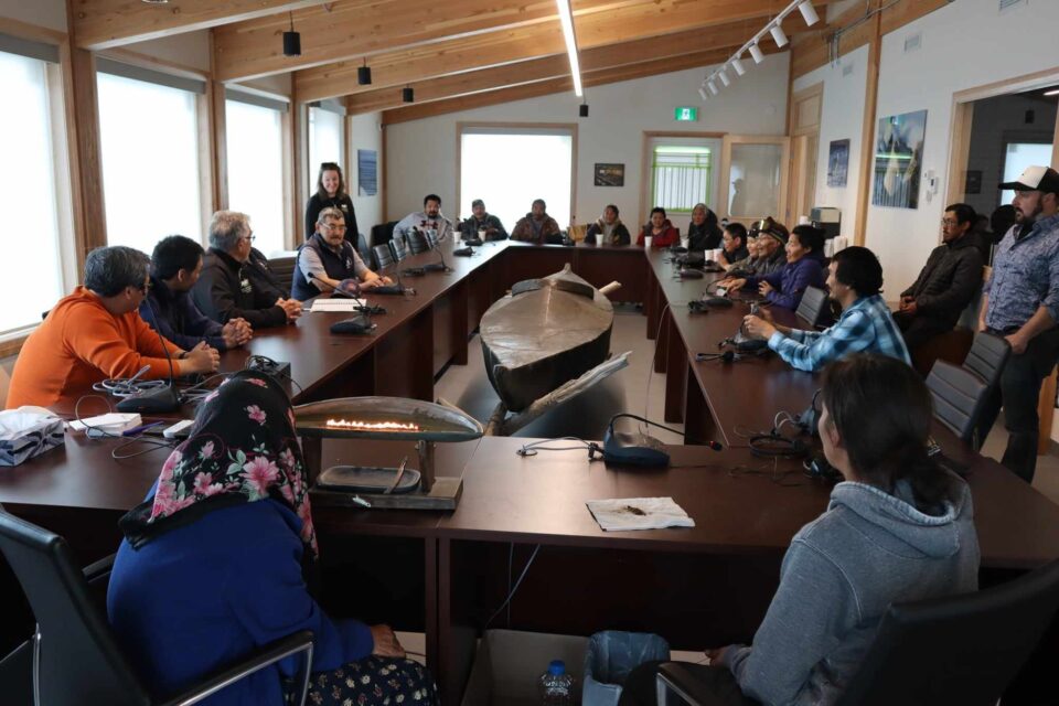 Diverse group of people seated around a long conference table, with a large traditional boat centrally placed at the Inuit consultation that led to signing of the energy purchase agreement for the wind turbines.