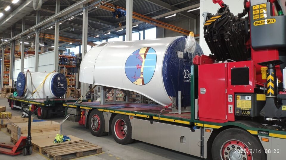 Large, white, cylindrical turbine parts with graphic design, secured on a flatbed truck, being transported in a factory setting.