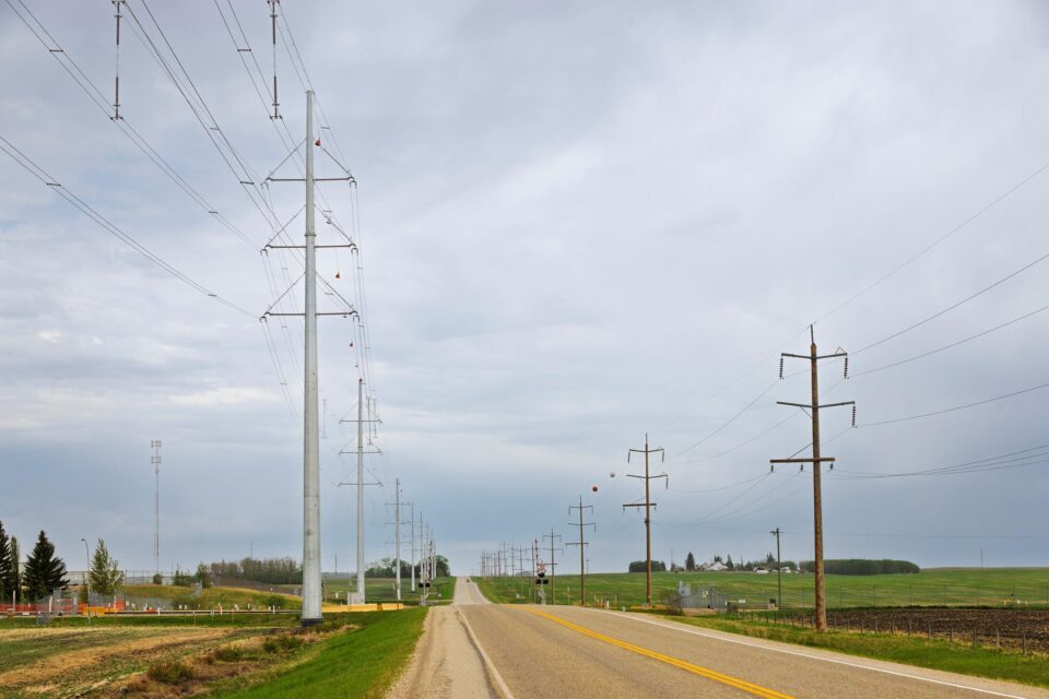 Long row of transmission towers and voltage lines along a paved road in a rural area.