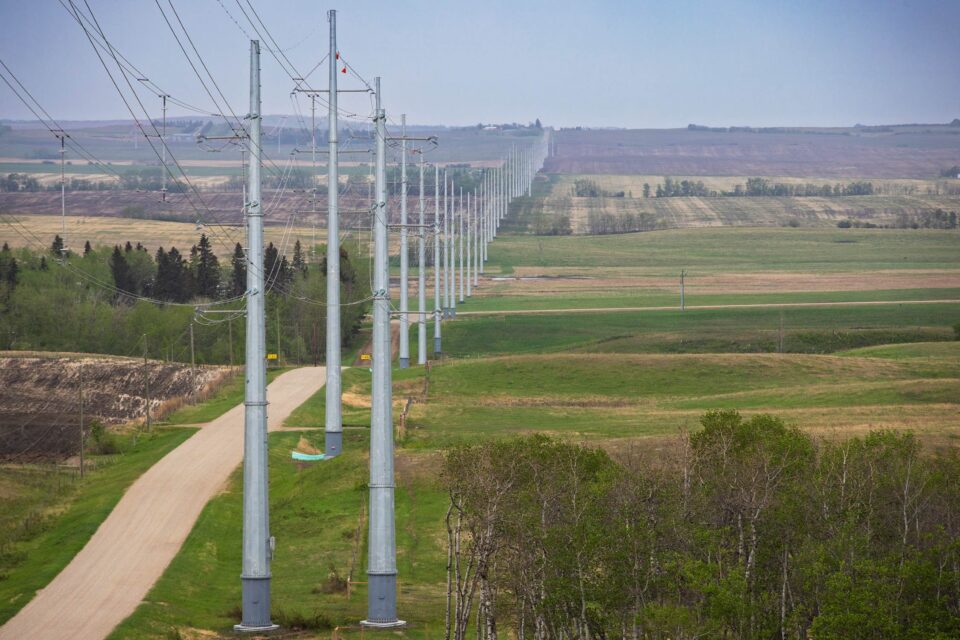 Metal transmission poles in the foreground and horizon that follows a dirt road and green fields.