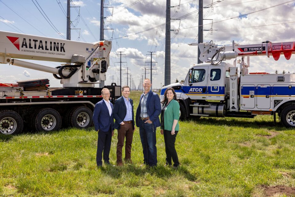 Four people stand together on a grassy field in front of two large utility bucket trucks. The trucks are branded “AltaLink,” and “ATCO” with “Terex” visible. Multiple tall electrical transmission towers and power lines extend into the background under a partly cloudy sky.