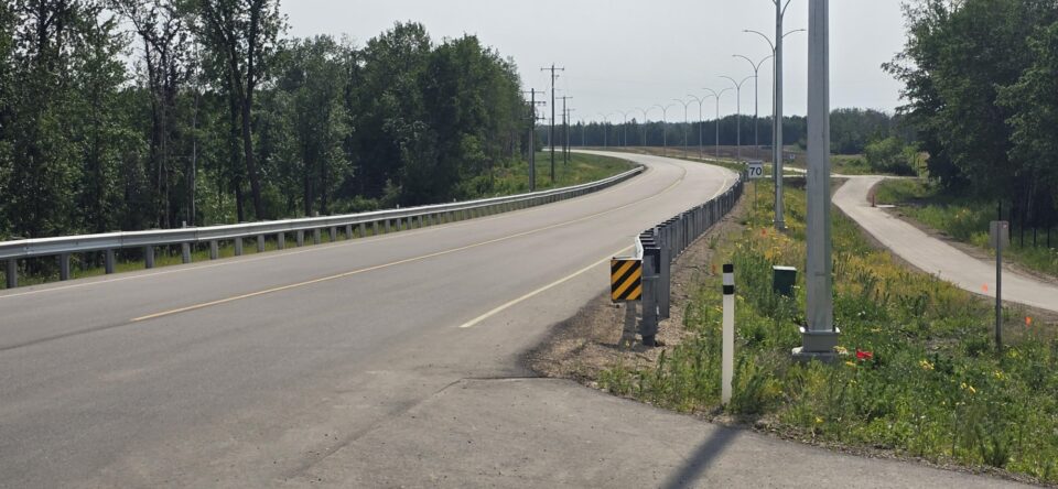 Route Enoch achevée : Une route à deux voies qui tourne vers la gauche avec une route secondaire qui bifurque vers la droite, entourée d’arbres et d’un panneau de limite de vitesse de 70 km/h.
