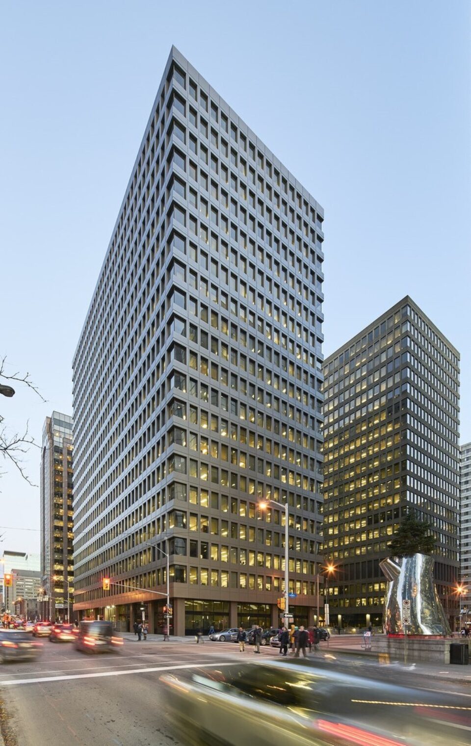 Deux tours de bureaux de hauteur moyenne à élevée avec des façades vitrées en forme de grille se dressent à une intersection du centre-ville au crépuscule, avec de nombreuses lumières intérieures visibles. Au premier plan, les voitures créent un flou de bougé en passant le long de la rue, tandis que les piétons se rassemblent près du coin. Une sculpture abstraite moderne trône sur le trottoir à droite, et les bâtiments et lampadaires environnants encadrent la scène urbaine sous un ciel pâle le soir.